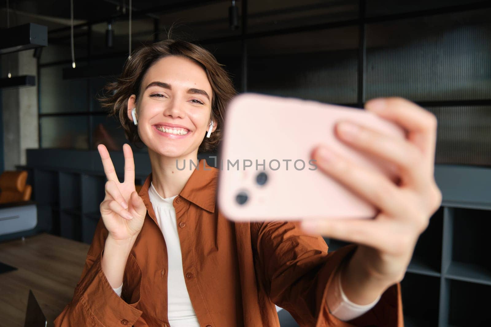 Portrait of beautiful smiling woman, taking selfie on smartphone, connects to online streaming, creating content for company, showing peace sign.