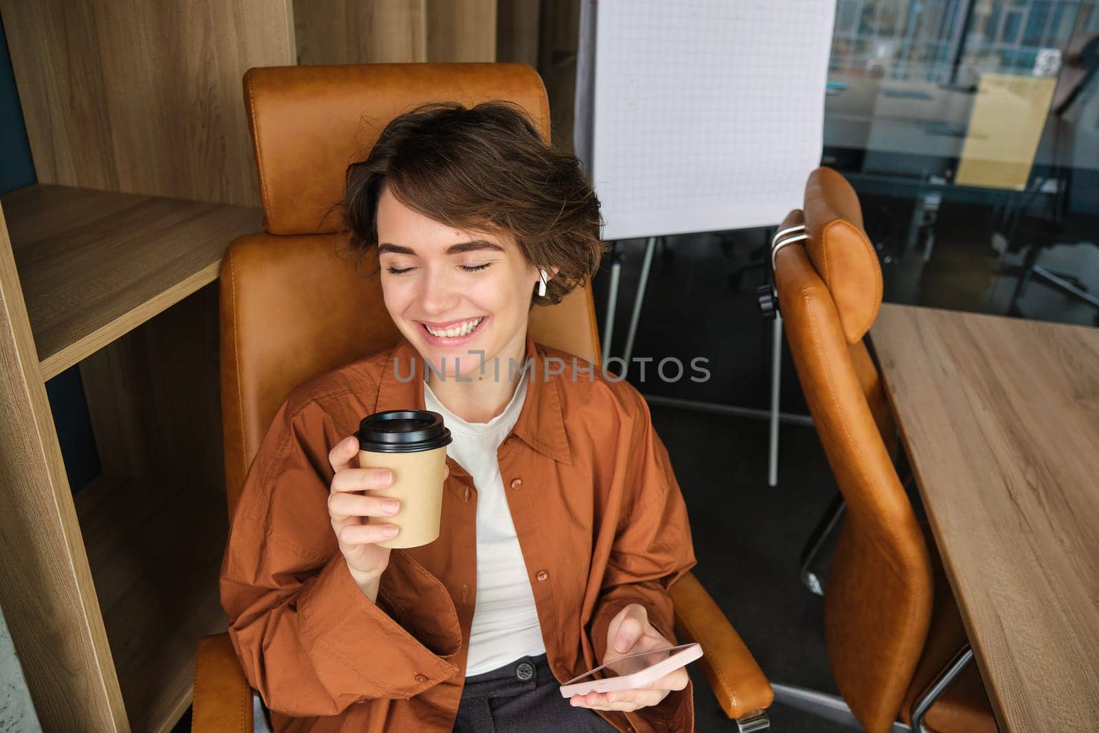 Portrait of happy young woman working in her office, sitting in chair with smartphone and coffee, drinking beverage from takeaway cup and smiling.