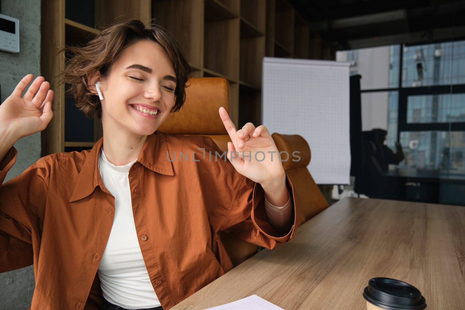 Portrait of young office employee, woman working in conference room, dancing while sitting with wireless headphones, listening to music.