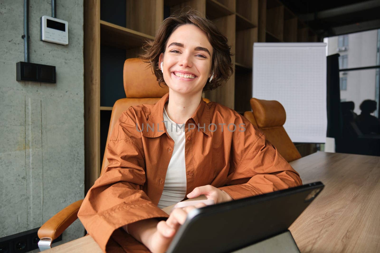 Portrait of young confident woman, employer in casual clothes, working in office, has digital tablet and coffee on tablet, smiling with self-assured face expression.
