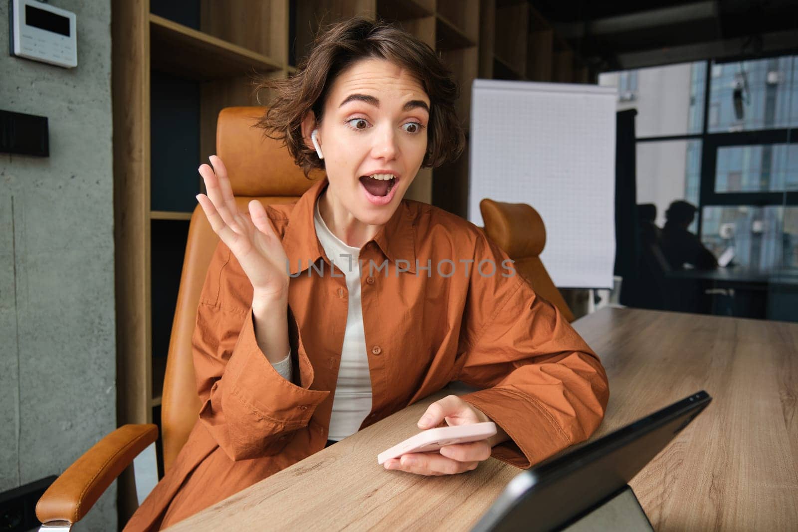 Portrait of excited young businesswoman, female entrepreneur in office, looking amazed, read great news on social media, holding smartphone and using digital tablet, enthusiastic reaction.