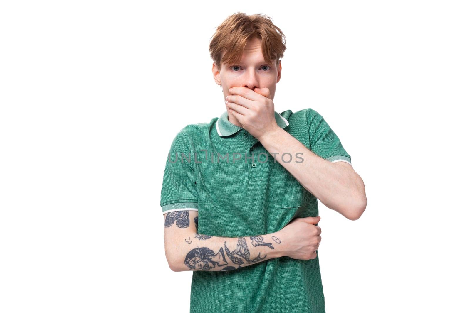 young slender ginger man with a tattoo on his arm dressed in a green T-shirt is shocked and covers his mouth with his hand.