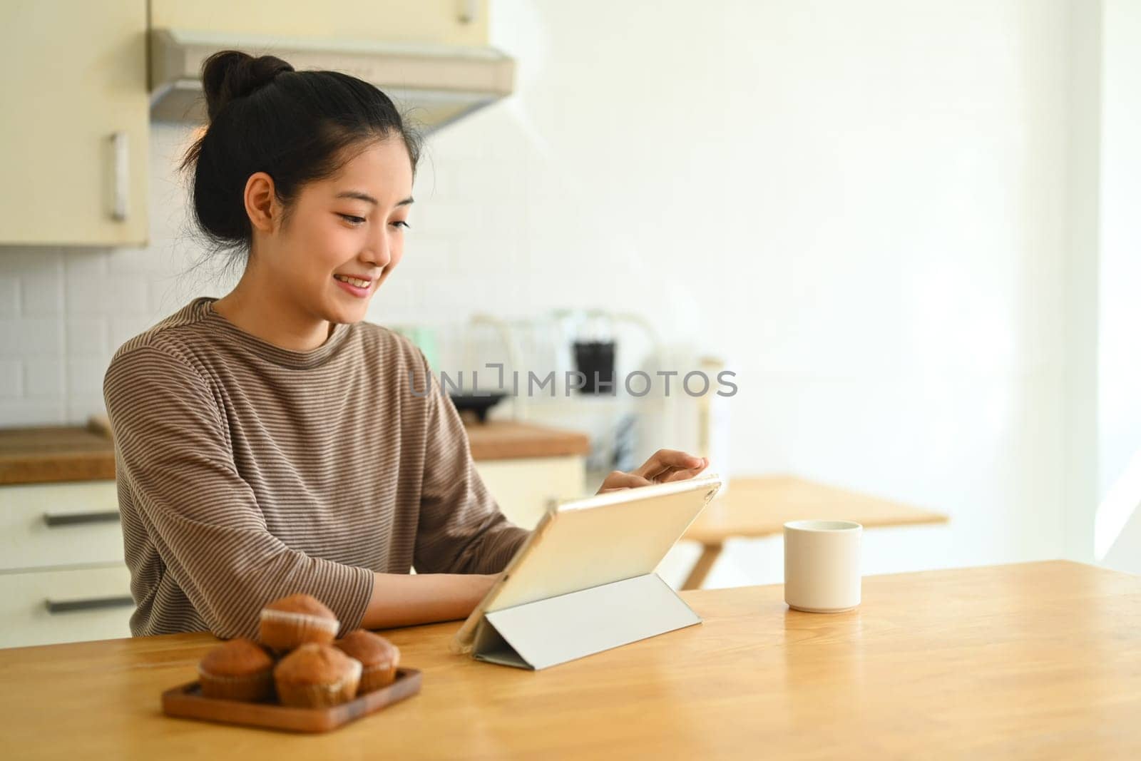 Young Asian woman sitting at kitchen table reading news or checking email on digital tablet.