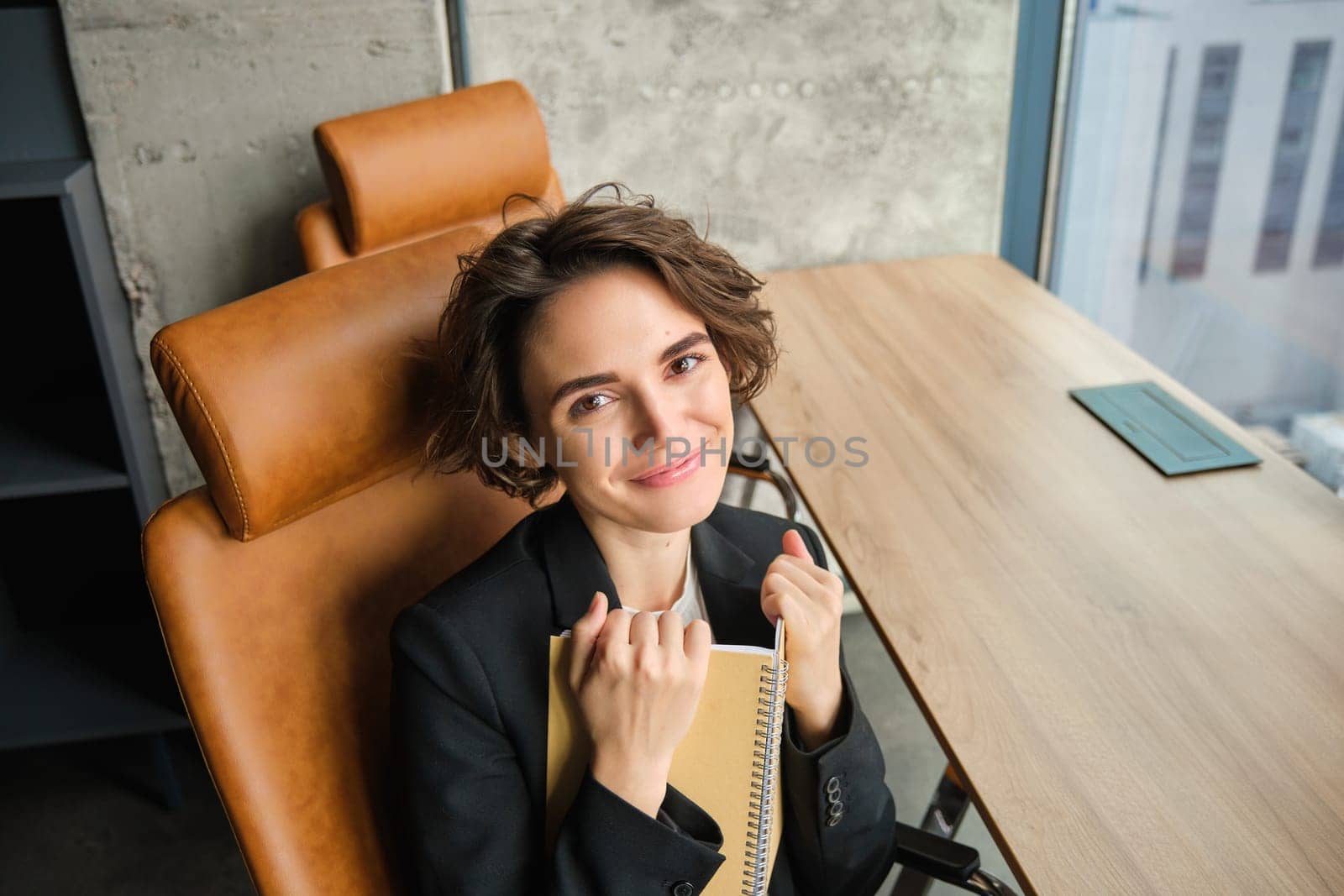 Top angle view of successful businesswoman, writing down information in notebook, working on planning her schedule, sitting in office and smiling at camera.