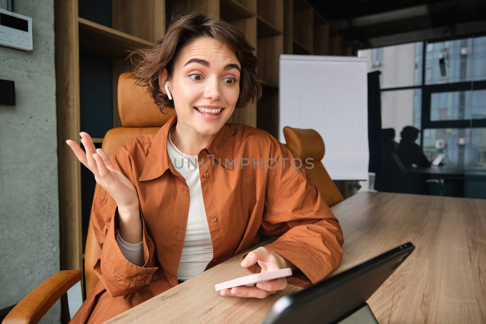 Portrait of woman working in office, looking amazed, makes surprised reaction to big news, holding smartphone, using digital tablet.