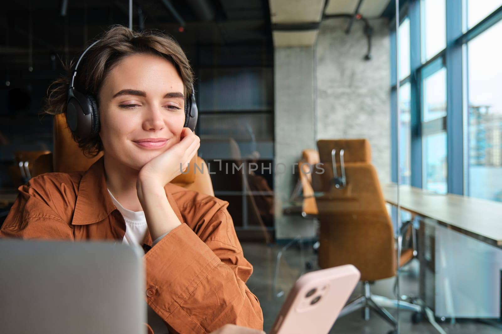 Portrait of woman checking her messages on smartphone, sitting in office with laptop, listening music in wireless headphones.