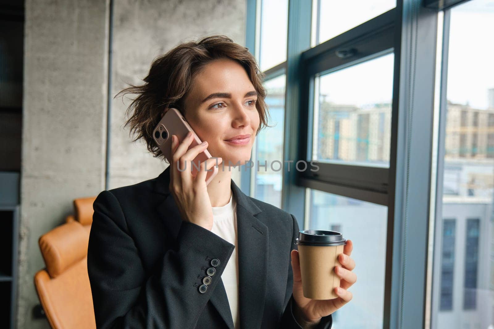 Image of female entrepreneur answering a phone call, standing in office and looking outside window, drinking coffee and having a conversation, wearing business suit.