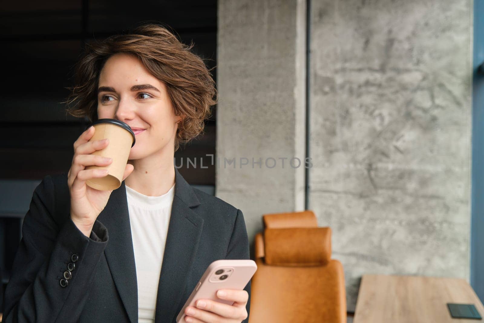 Image of successful female entrepreneur, businesswoman drinks coffee in her office and holds mobile phone.