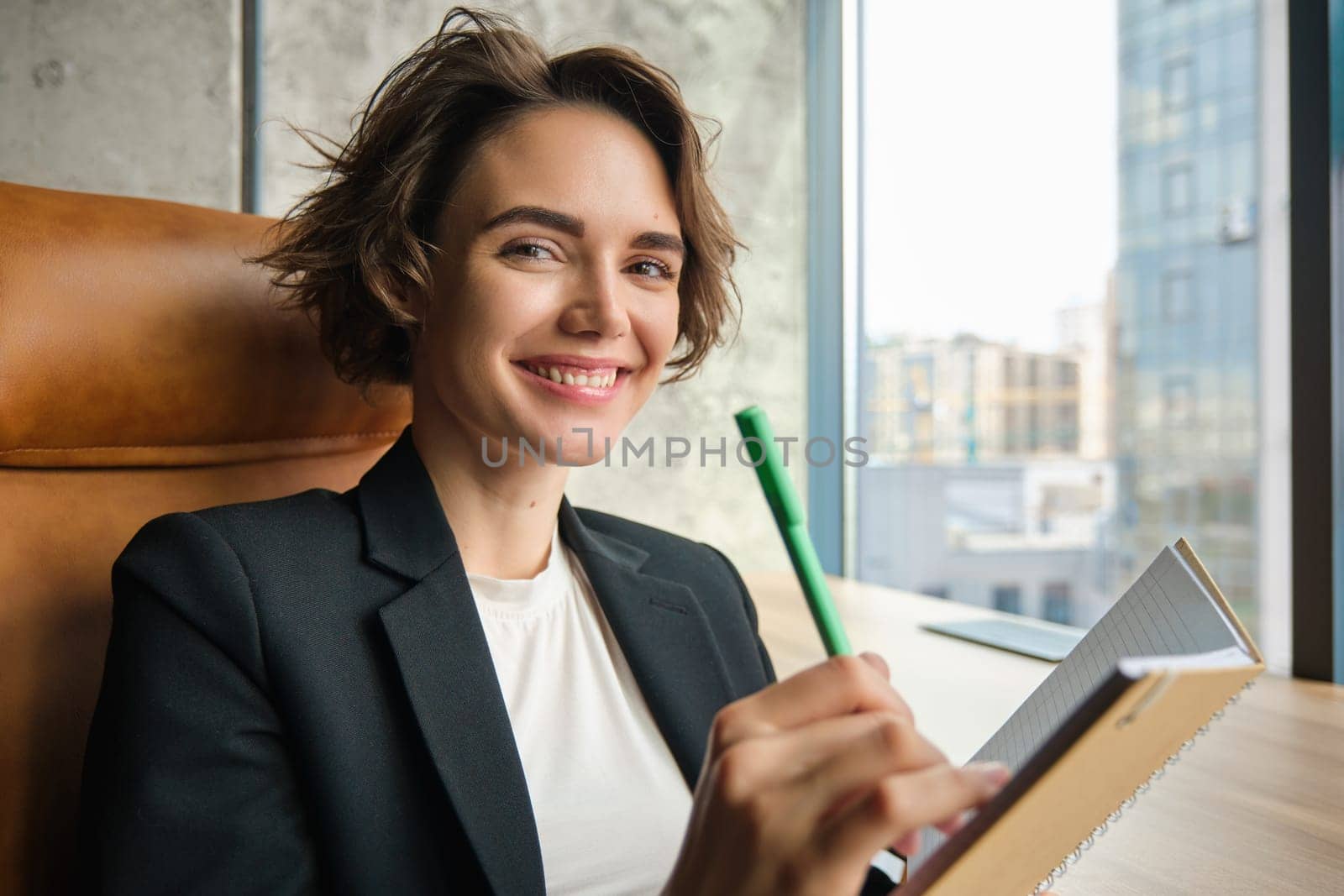Portrait of confident saleswoman working in an office, sitting in business room, reading, writing down her thoughts for presentation, making notes, sitting near window, preparing for meeting.