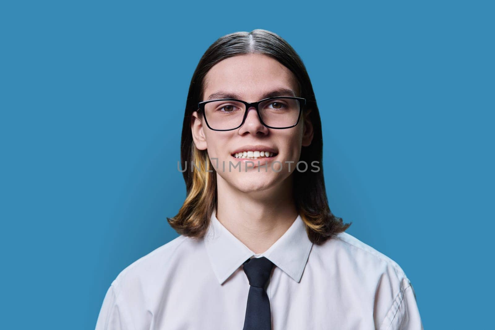Headshot portrait of smiling teenage guy 18, 19 years old, on blue color studio background. Positive handsome young male in glasses shirt and tie looking at camera