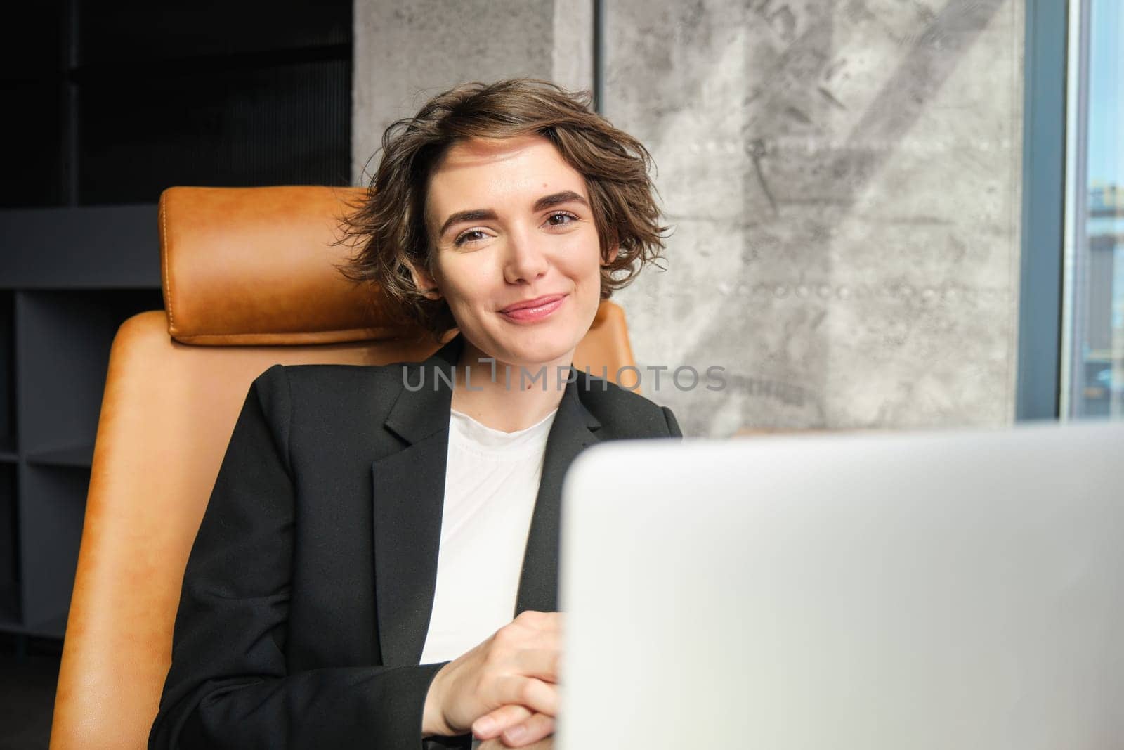 Portrait of confident, successful businesswoman in suit, sits in her office with laptop, working on project, watching webinar, being present at online conference or meeting.