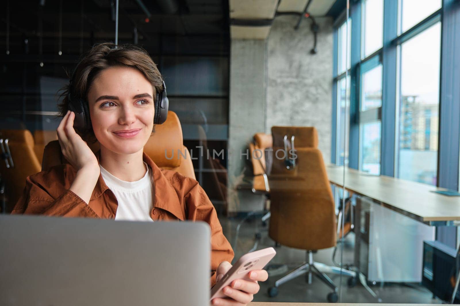Close up portrait of working woman, sitting in office with laptop, connects to video chat in headphones, holding smartphone, joins online meeting.