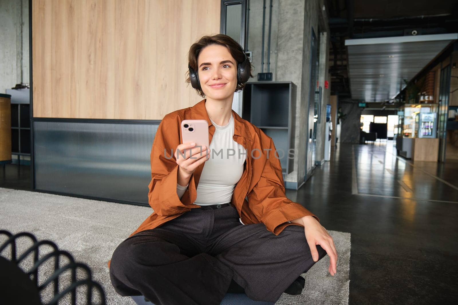 Portrait of happy young woman, manager in office, sitting in lounge room, relaxing on lunch in break room, holding smartphone, listening music in wireless headphones, using co-working space.
