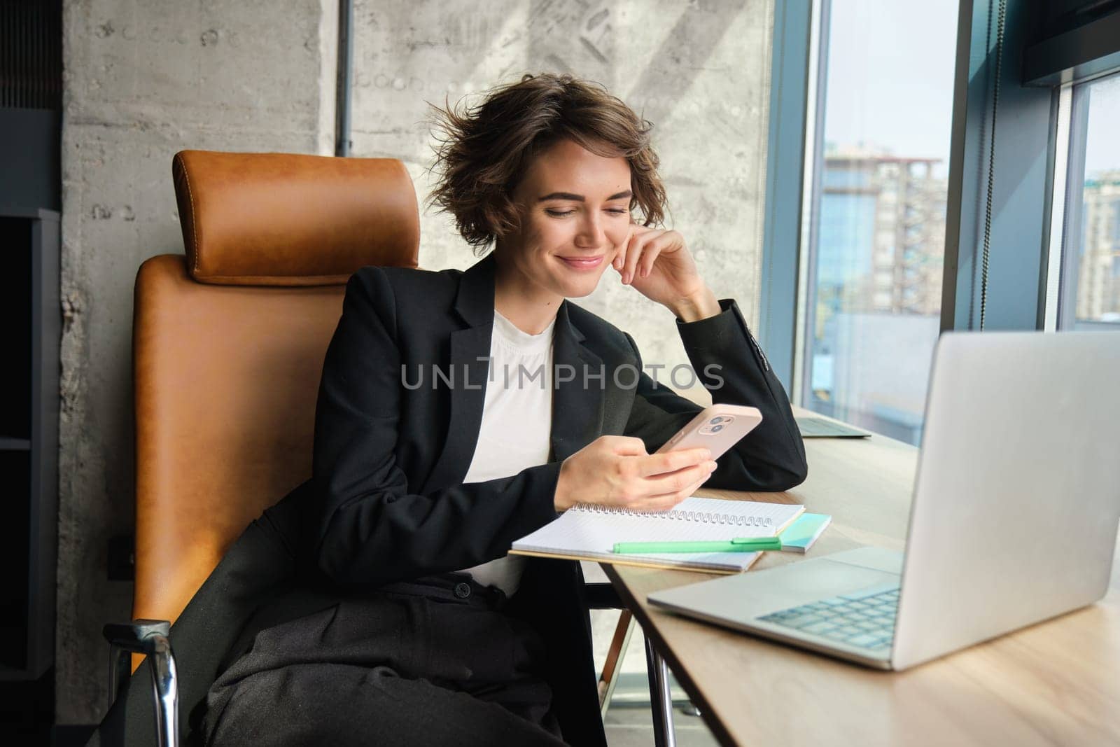 Portrait of woman in an office, working on laptop, making notes, sitting near window.