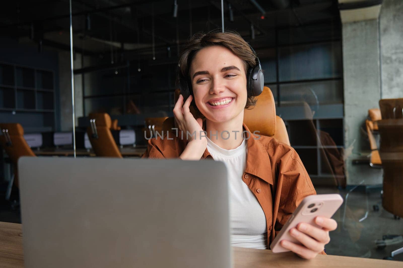 Successful business. Smiling woman entrepreneur, digital nomad in headphones, sitting in office with laptop and smartphone, working at workplace.