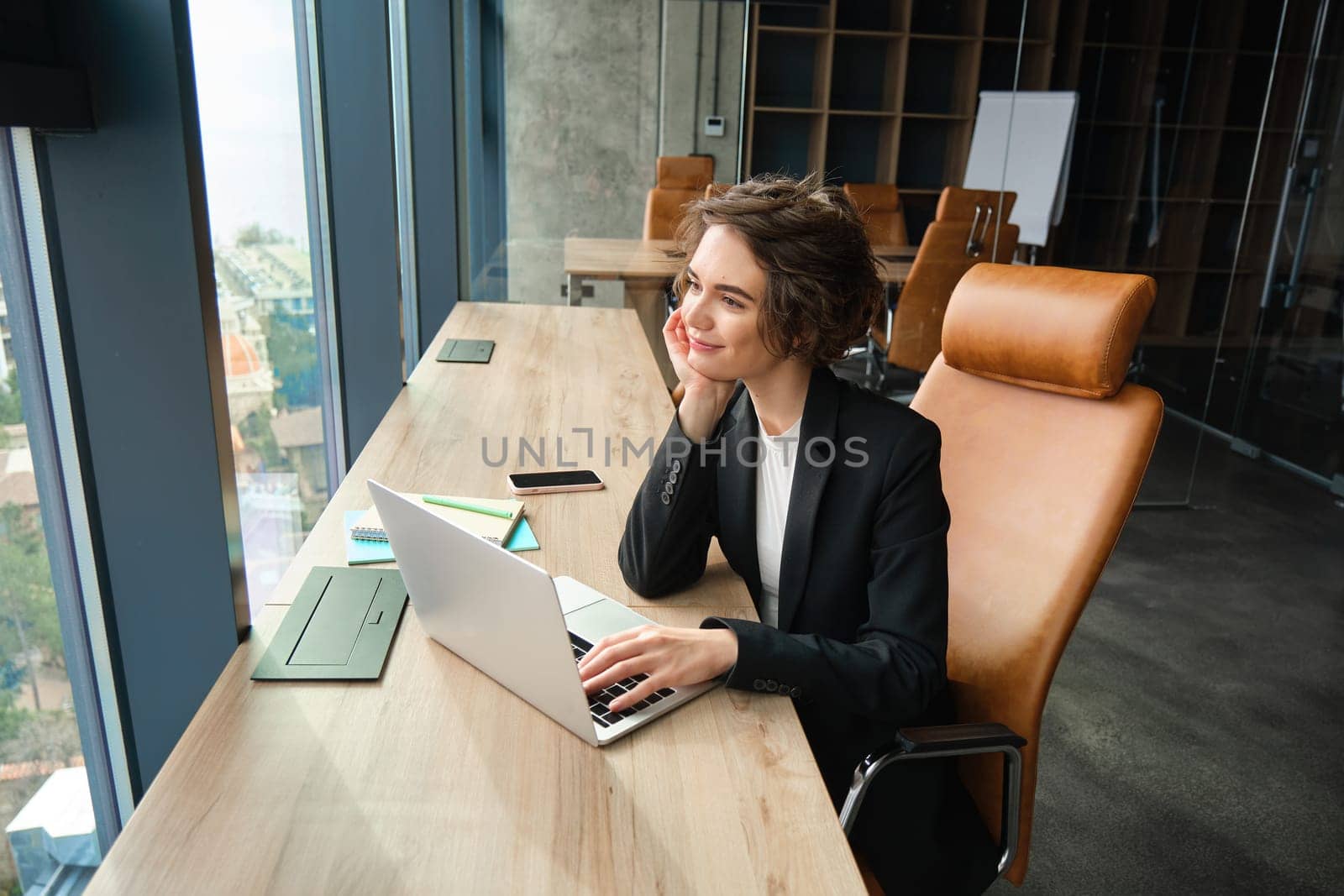 Businesswoman working on laptop, sitting in office in suit, waiting for client, preparing documentation for corporate meeting, using coworking space to focus on her work.