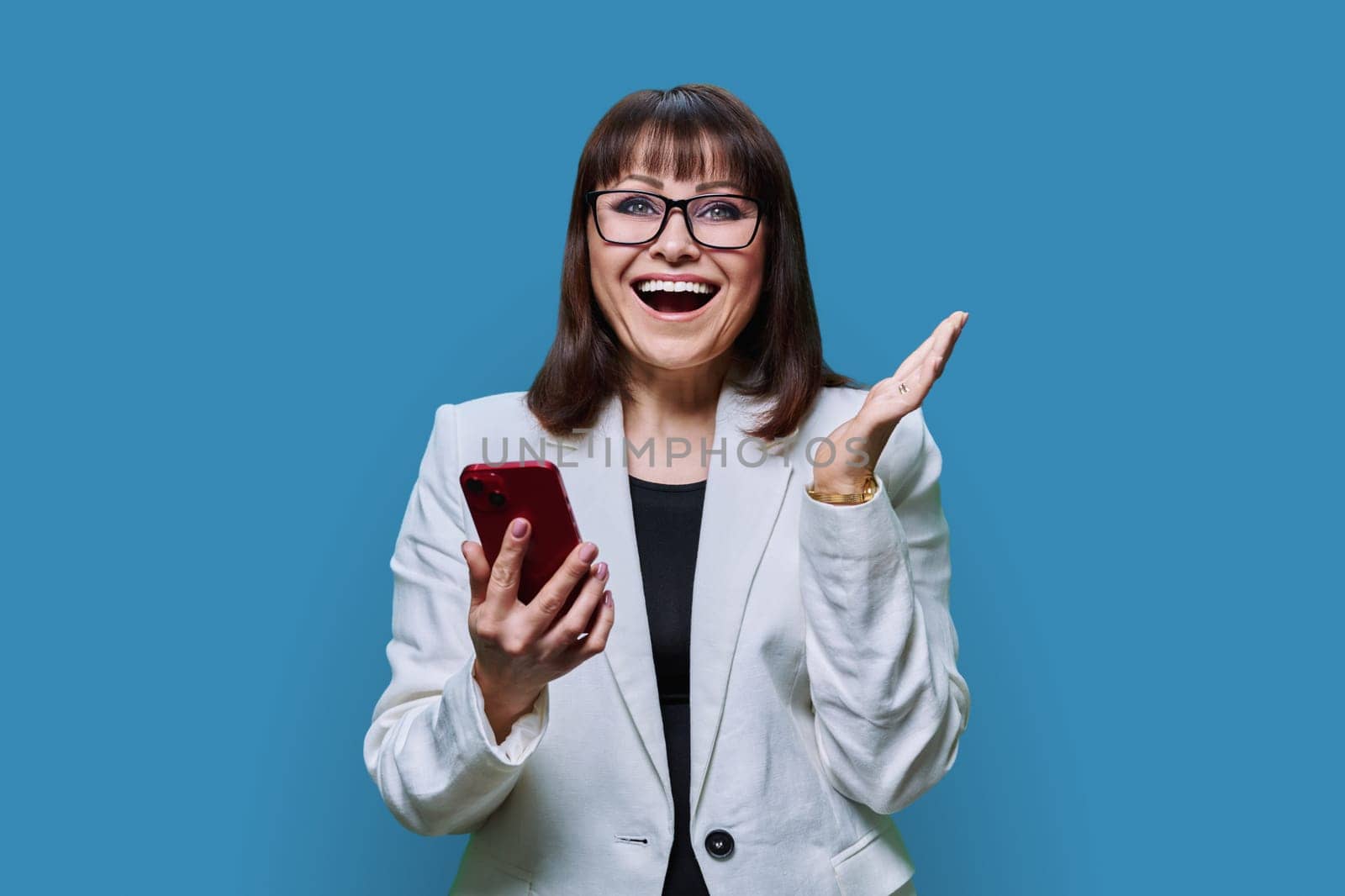 Portrait of surprised middle aged beautiful businesswoman with phone in hands on blue studio background. Smiling mature female in jacket, glasses looking at camera. Technology, business, people