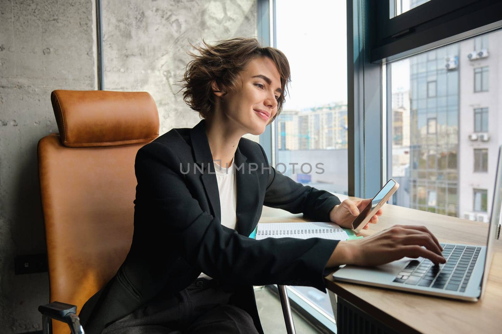 Young working woman in office, sitting in front of computer and using smartphone, wearing suit.