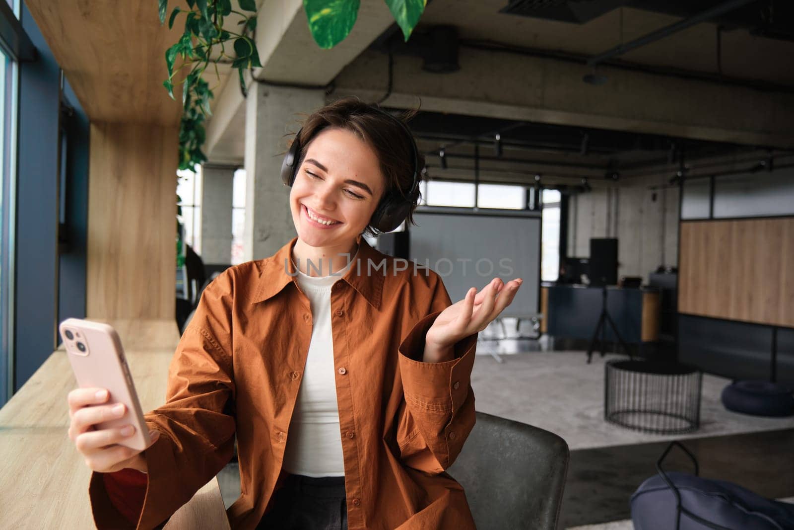Image of smiling young woman, company employee, sitting in office with smartphone, demonstrating creative co-working space to people online, showing smth, video chats on mobile phone.