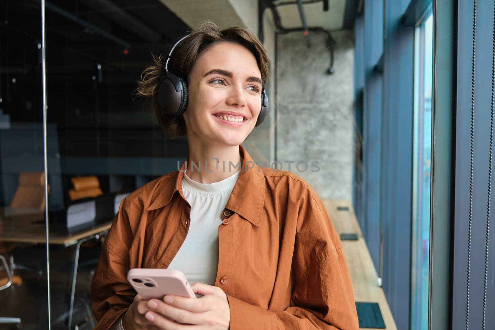 Portrait of woman working in office, digital nomad in casual clothes, wearing wireless headphones, holding mobile phone, looking outside window with pleased smile.