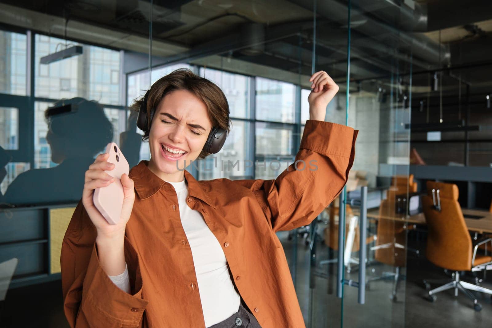 Business and success concept. Happy young woman dancing in her office, employee having fun, listening music in headphones, holding mobile phone.