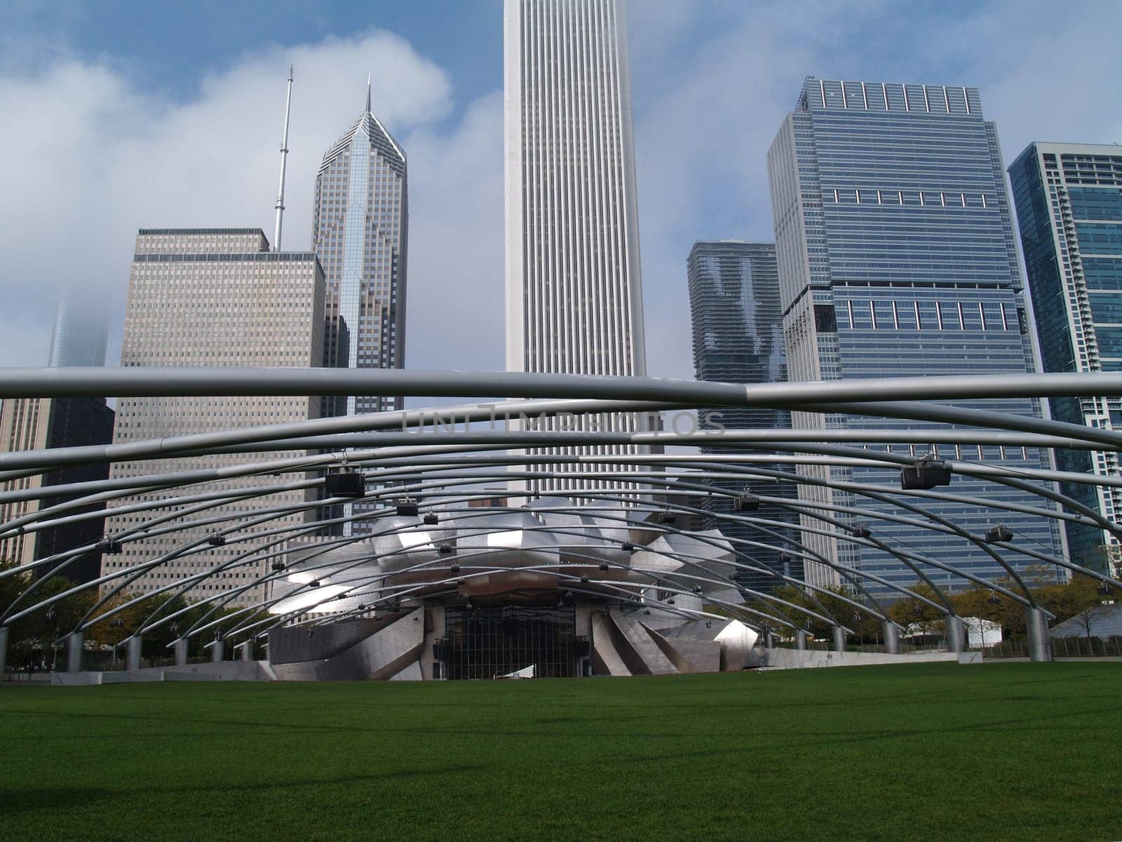 Grassy seating area of the Jay Pritzker Pavilion  showing the sound system supported on trellis style pipes in front of a Chicago skyline in Millennium Park.