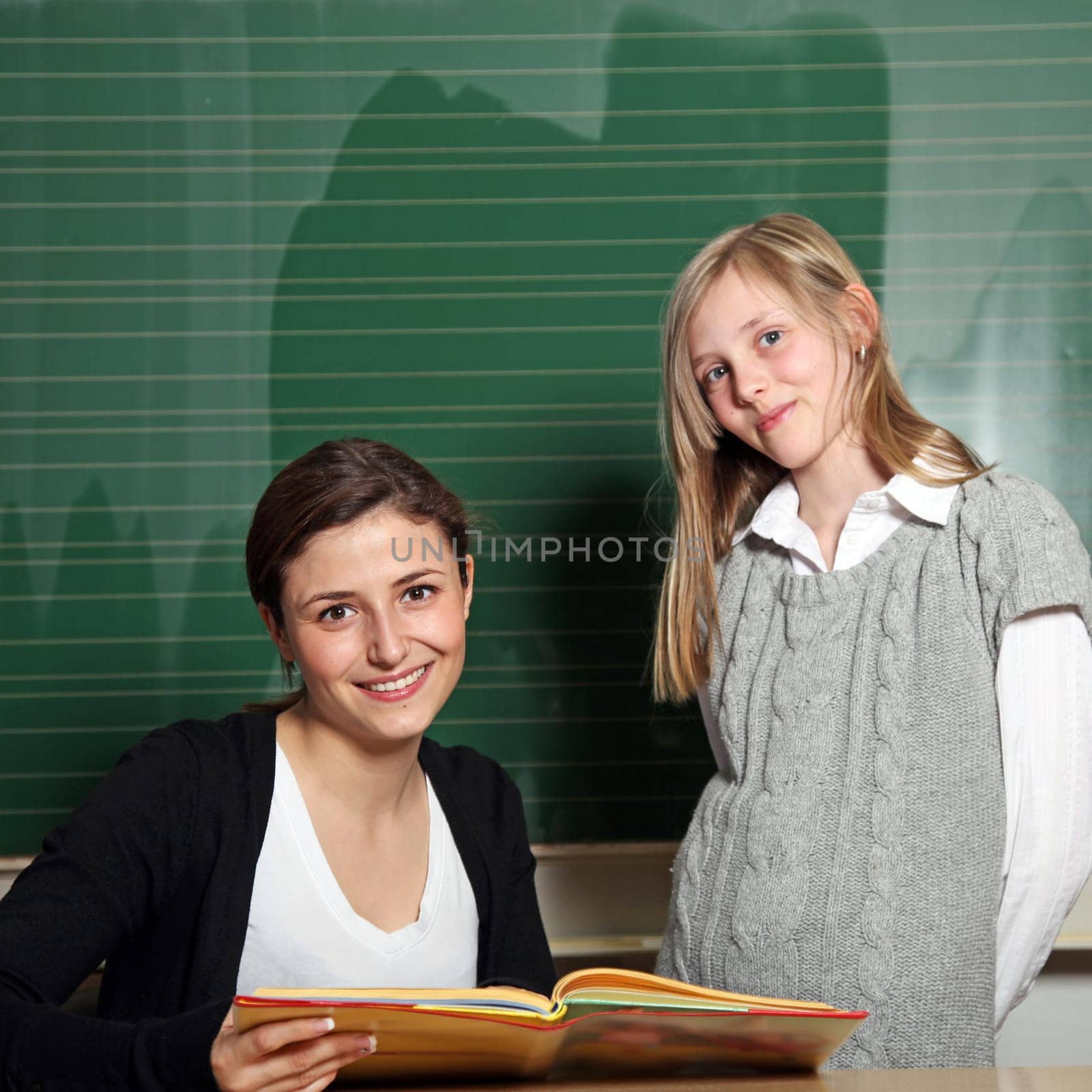 Cute teacher with textbook and pupil in front of the blackboard. Both smile at the camera