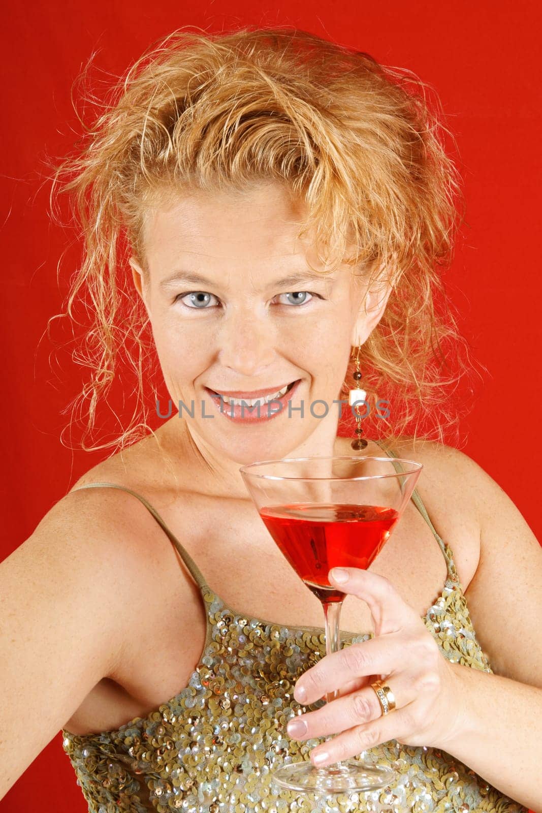 Portrait of a beautiful blond woman with blue eyes, wearing a green sequins top, holding a red drink, over a red background.