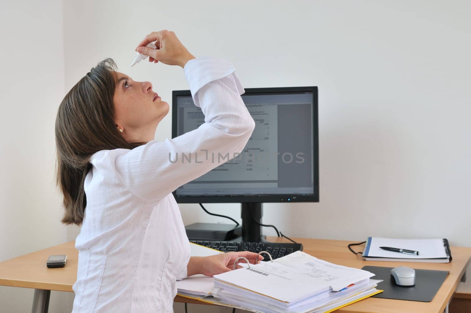 Detail of young business person (woman) applying eye drops on workplace - computer on table