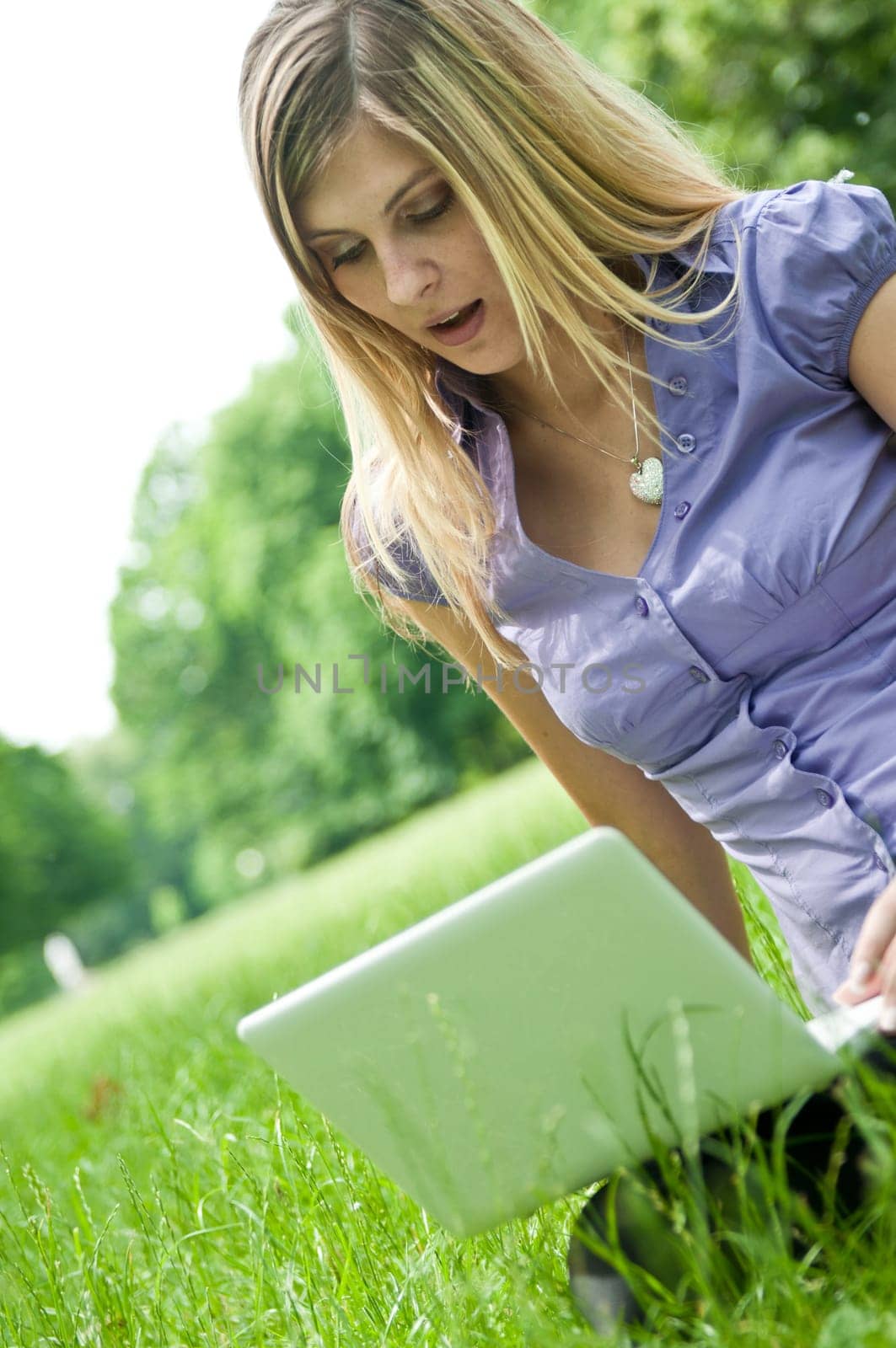 Young blond woman working with netbook outdoors in park on grass - surprise expression