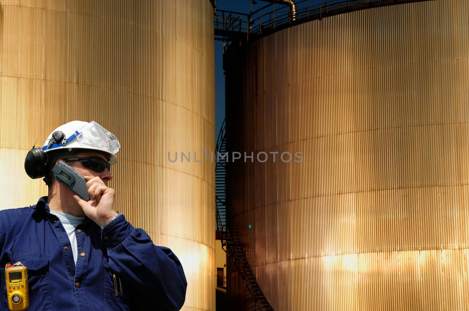 engineer, worker in hardhat, in front of sunlit large fuel-storage towers, tanks, inside oil and gas refinery