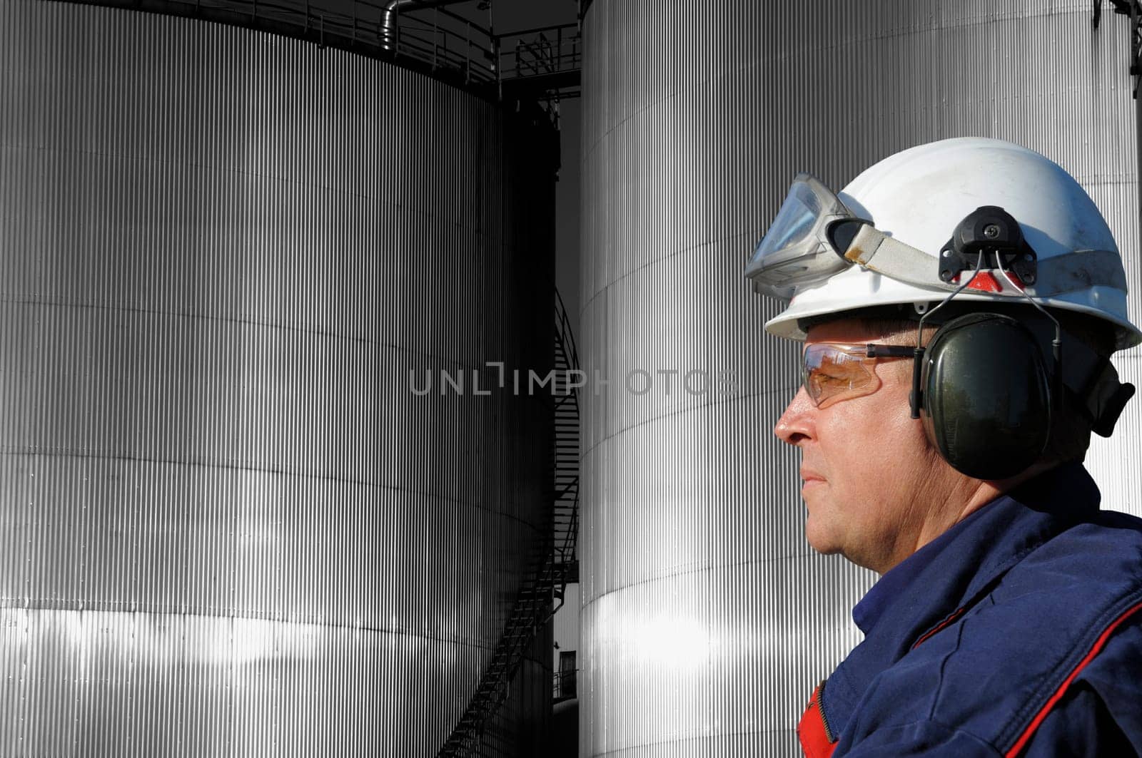 engineer, in profile, large industrial fuel-storage tanks in background, sunlight hitting the metal