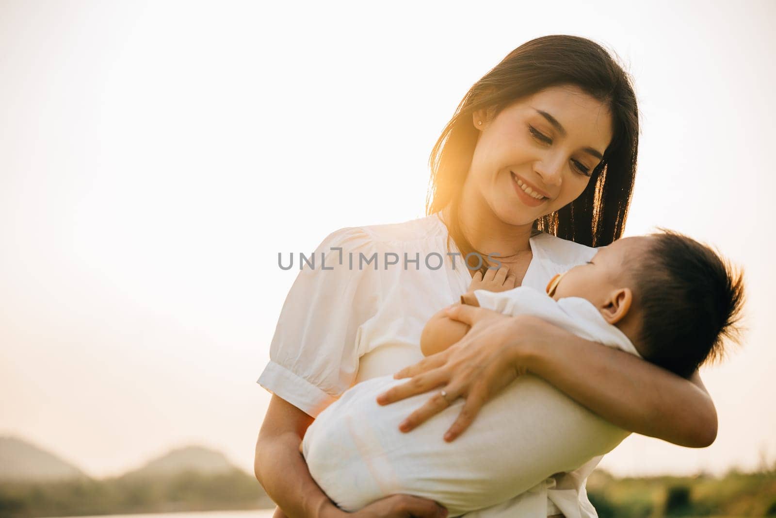 Family care. Happy beautiful mother holding baby sleeping son in the park at evening time before sunset, Portrait of mom hold newborn feeling happy and smiles in the garden nature outdoor, Mother Day
