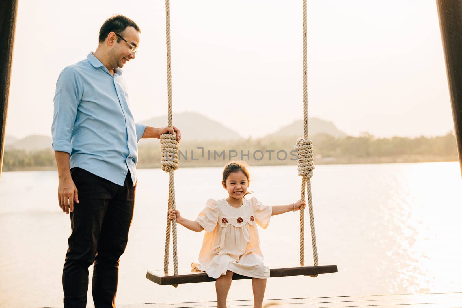 A happy Asian family spending quality time together on a sunny summer day at the playground, with the father pushing his cute daughter on a swing and both of them smiling and laughing with joy