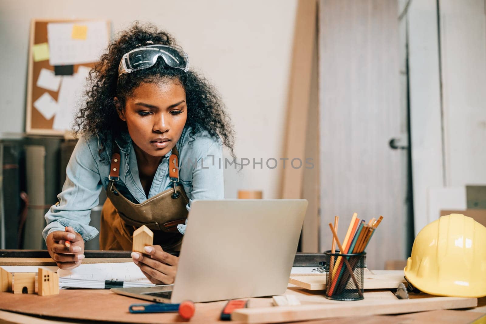 Carpenter america black woman curly hair sketch making notes in work paper while standing at wooden table with laptop computer, young female working learning online at woodshop, Happy Carpenters Day