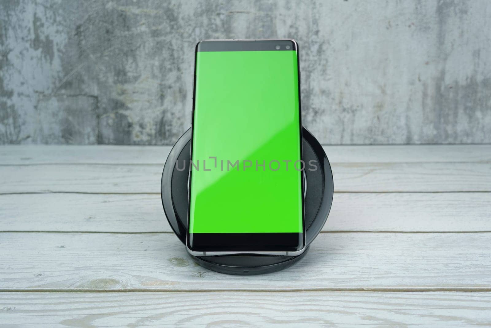 Modern smartphone with a bright green screen placed on a wireless charging pad on a wooden surface, with a textured gray background, ideal for mockups and design projects.