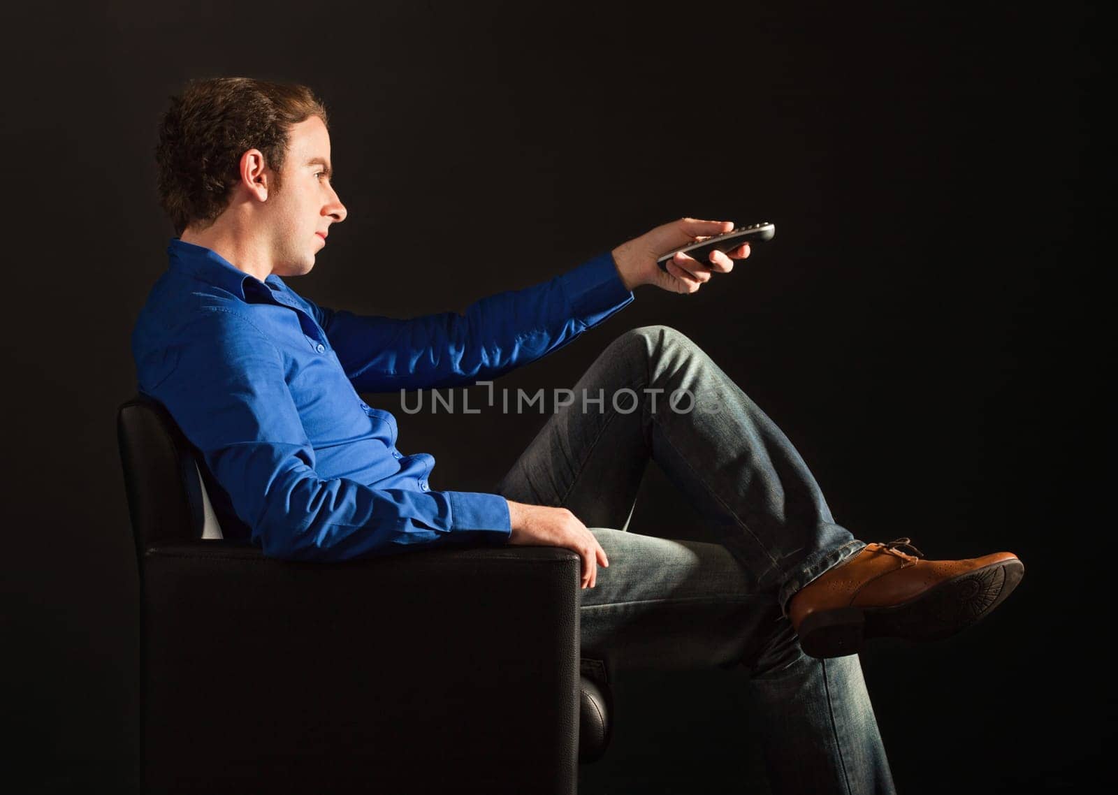 Handsome man sitting on the armchair in the dark changing TV channeels with remote control