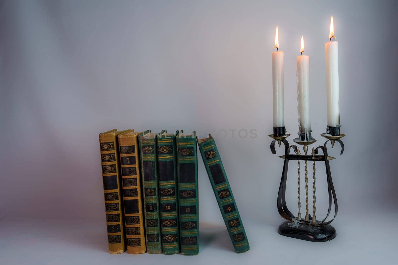 A row of antique hardcover books leaning slightly, next to a vintage candelabrum with burning candles, creating a nostalgic and moody atmosphere