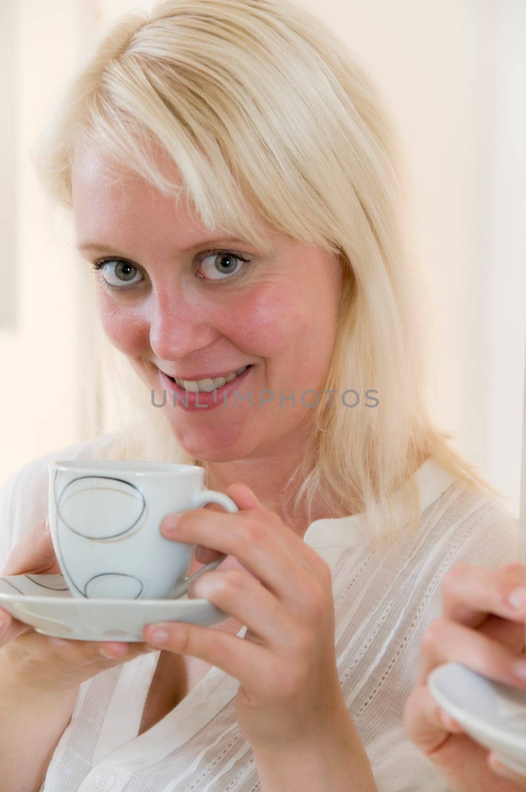 Portrait of young blonde woman holding coffee cup in front of bright kitchen in background and looking at camera