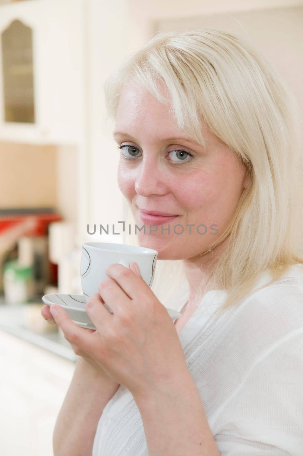 Portrait of young blonde woman holding coffee cup in front of bright kitchen in background and looking at camera