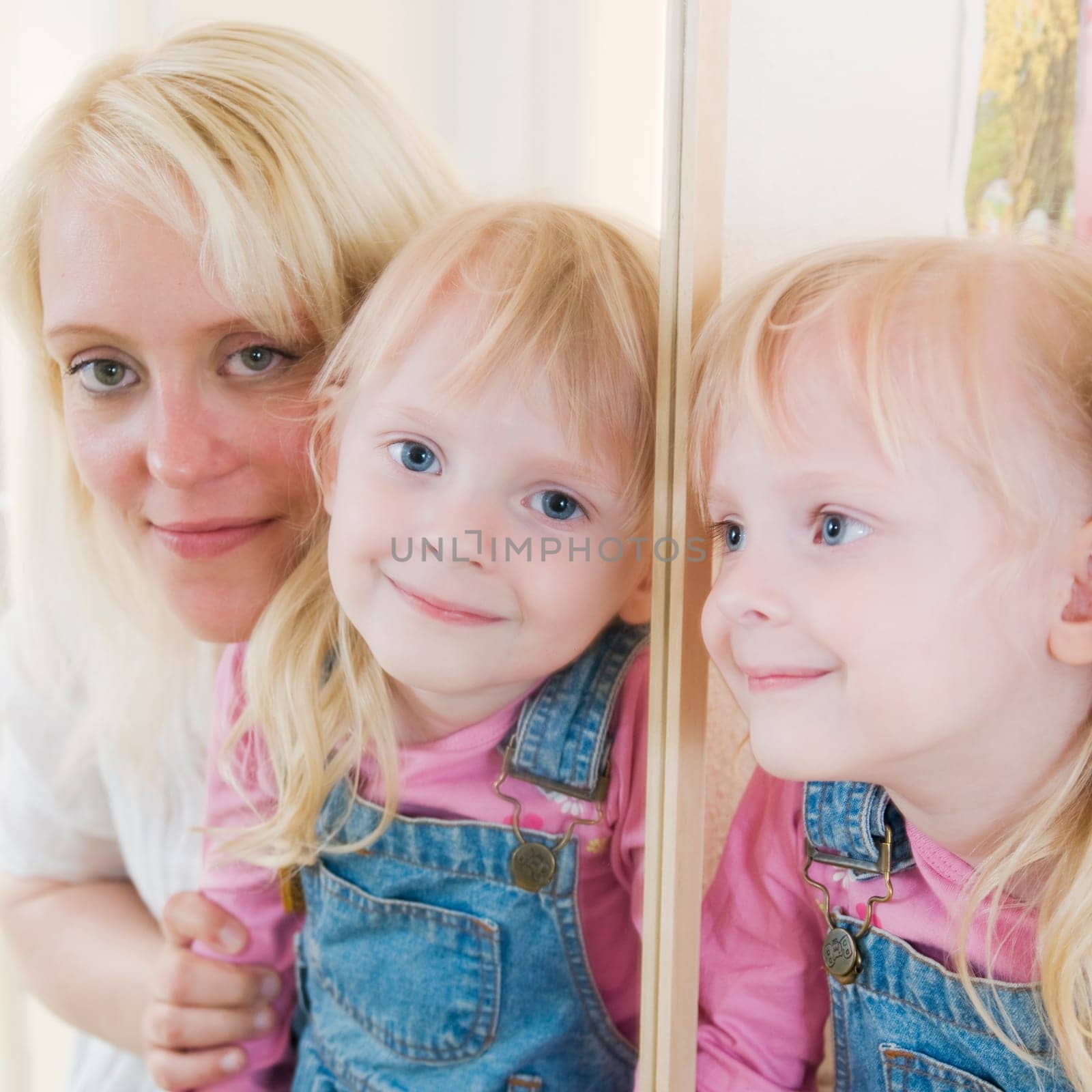 Portrait of a young blonde mother with her blonde four-year-old daughter in the mirror image looking at the camera. Daughter is originally depicted looking to the side and mirror-image.