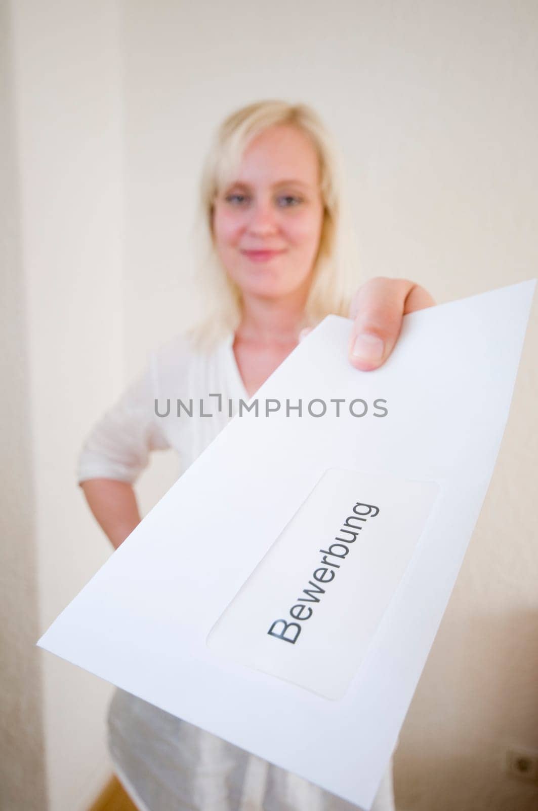 Young blonde woman handing over her application letter in an envelope in front of a white wall towards the camera