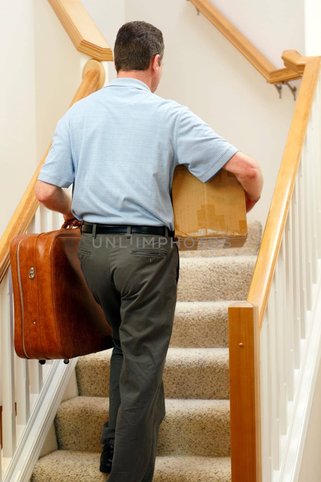 Man dressed in business casual clothes walking up stairs with suitcase in one hand and box under right arm.