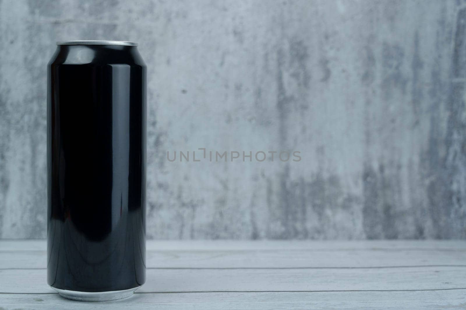 Blank black aluminum can standing on a wooden surface against a gray textured wall, suitable for branding and product mockup.