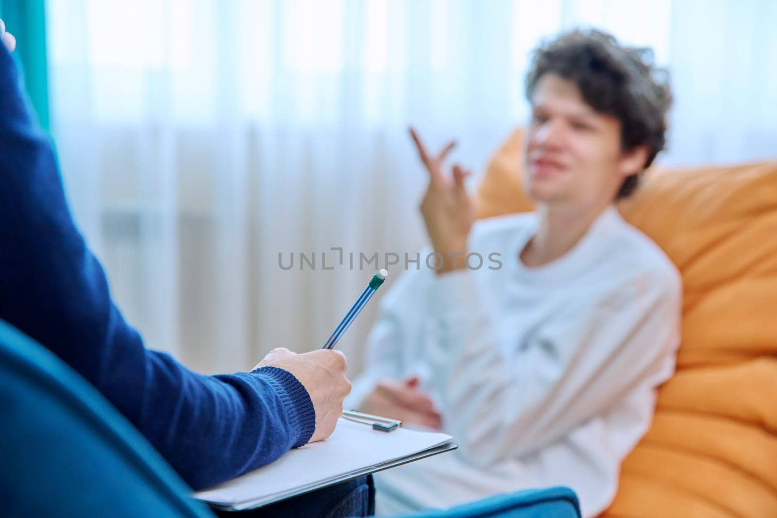 Close-up of female psychologist's hands with clipboard making notes, in individual therapy session. Young male patient in chair out of focus. Psychology psychotherapy treatment mental health concept