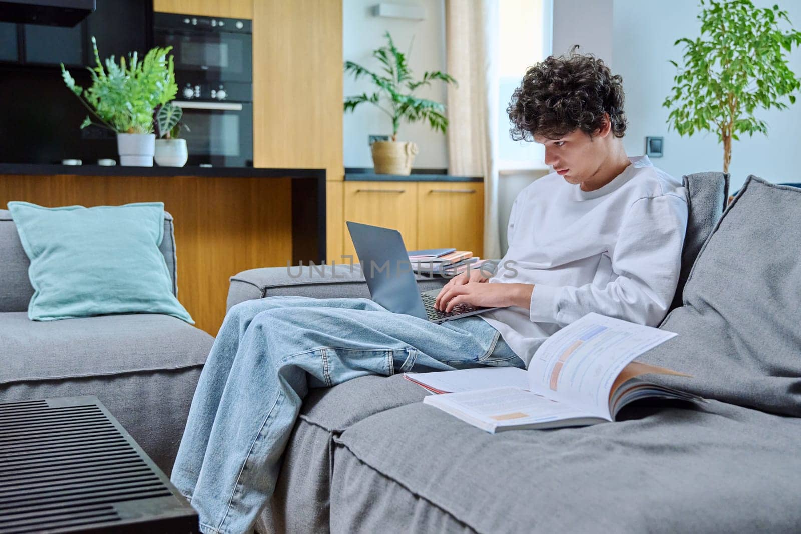 Handsome guy college student studying at home, sitting on sofa typing on laptop. Young male 18, 19 years old with curly hair in interior of living room. Education knowledge online internet technology