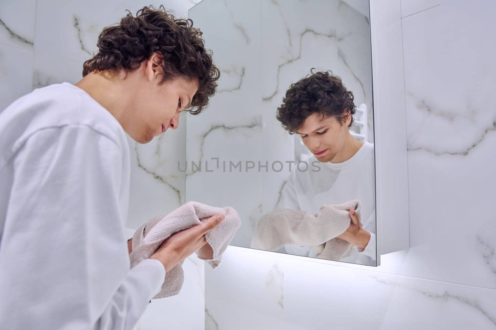 Handsome young guy 18-20 years old in bathroom near mirror, washing his face, wiping with towel. Morning evening routine, hygiene, skin care facial cosmetics for young males