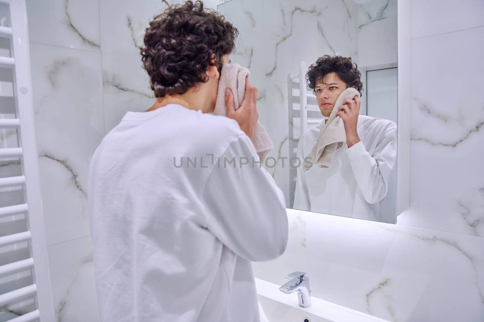 Handsome young guy 18-20 years old in bathroom with towel looking at his reflection in mirror. Morning evening routine, hygiene, skin care facial cosmetics for young males