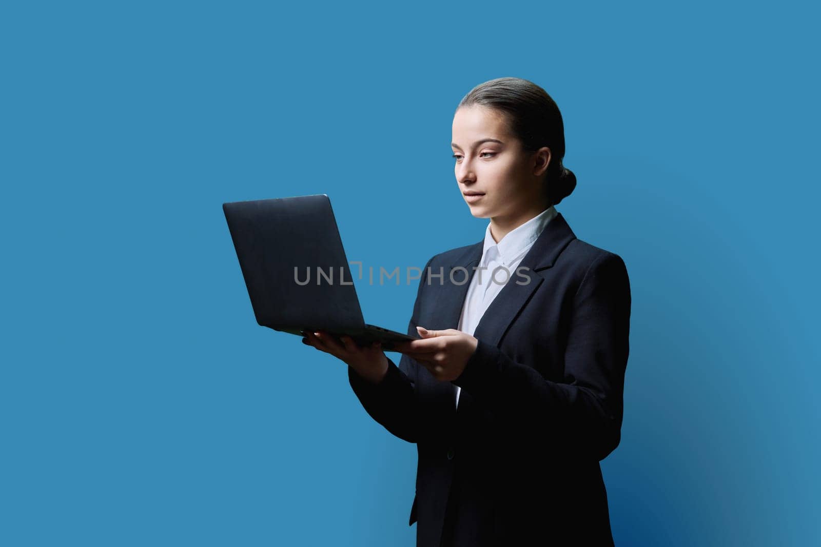 Serious teenage female student in black jacket using laptop on blue studio background. Education, learning, technology, knowledge concept