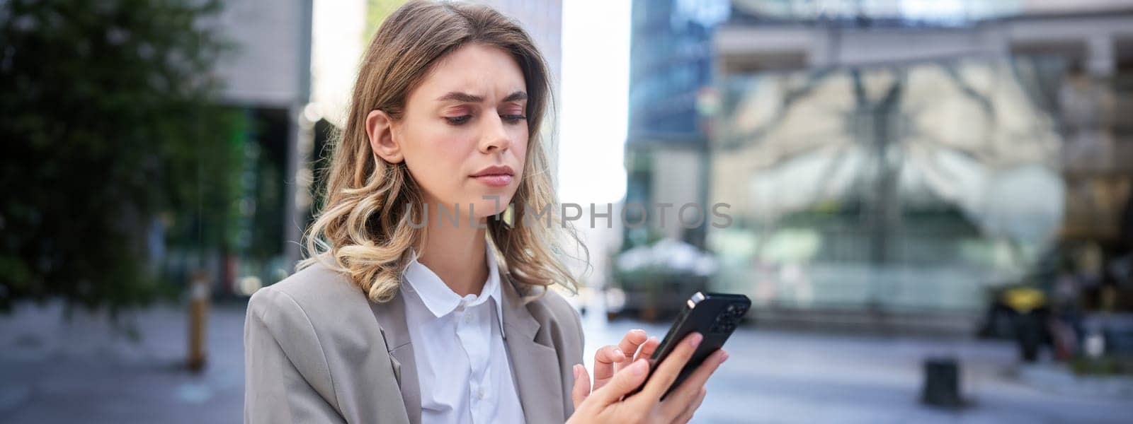 Young businesswoman looking complicated, frowning as reading message on mobile phone. Serious corporate woman checking her smartphone by Benzoix
